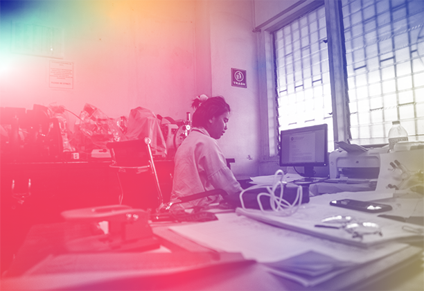 A person sits and works at a cluttered desk in a room filled with office and lab equipment, looking at papers and a computer screen. The grayscale image is overlaid with a multicoloured gradient filter, creating a vibrant, stylised atmosphere.